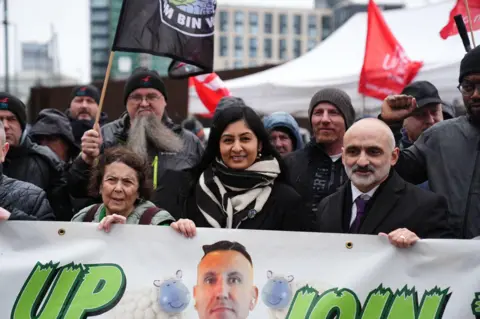 PA Media A smiling Zarah Sultana stands behind a banner. She is wearing a black and white scarf. To her left is Unite's national officer Onay Kasab. He has a grey beard and is wearing a white shirt, purple tie and black coat. 