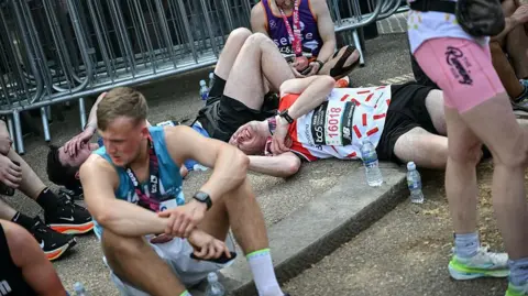 Getty Images Several marathon finishers sit or lie flat on the ground in a designated rest area, showing visible signs of fatigue after completing the race.