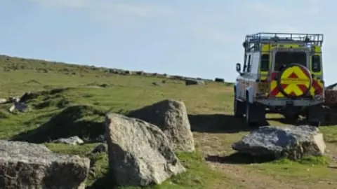 A view of a 4x4 on the right on the moor. It is yellow and red on the back and is parked on green moorland which has grey rocks scattered around.