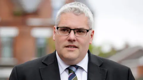 A close-up image of Gavin Robinson. He is a middle-age man with short, grey hair and is wearing a pair of dark, rectangle glasses. He has his mouth slightly ajar and is wearing a black suit jacket, a white shirt, and a colourful striped tie. He is standing near a red brick building, but it is blurred.