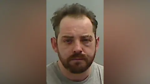 Mugshot of Sean Miller. He has a short ginger beard and short messy dark hair. He has a cut on his nose and swelling and bruising around his left eye.