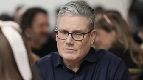 PA Sir Keir Starmer looks down as he sits at a table while wearing a navy shirt without a tie.