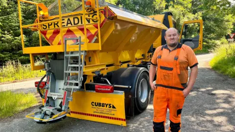 BBC Road worker in orange high-visibility clothing standing beside a yellow gritting truck marked Spreading on a rural roadside with trees in the background.