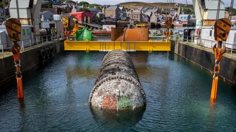 Microsoft A long, white cylinder covered in grime is being lifted by a crane from the sea. The Microsoft logo is just visible on the end cap beneath a layer of dirt