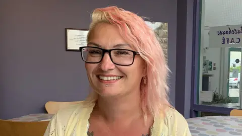 Charlotte is sitting in a cafe, wearing a yellow top. She has pink hair and wears glasses.