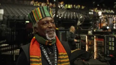 Liverpool poet Levi Tafari wears a hat and scarf in the colours of the Jamaican flag. He has grey hair and a grey beard.