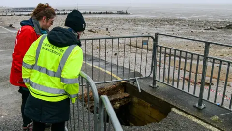 Borough Council of King's Lynn & West Norfolk Two women looking at a fenced-off sinkhole on the edge of a promenade. The women are on the left. Beyond the sea wall is a beach and the sea. 