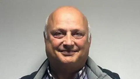 Russell Wate looks and smiles directly at the camera. He is a bald man with white hair on the sides of his head wearing a grey-and-white checked shirt and a dark top with a grey collar.