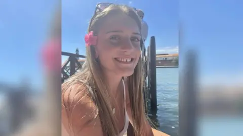 Family handout Juliette, smiling at the camera on a sunny day, is wearing her long hair down, sunglasses on her head and a pink flower tucked above her ear. She is sitting by the water