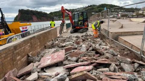 Construction workers in high-visibility clothing break up a concrete walkway beside a brick wall at a coastal site, using a small excavator and tools, with piles of rubble in the foreground and the sea and hillside buildings in the background.
