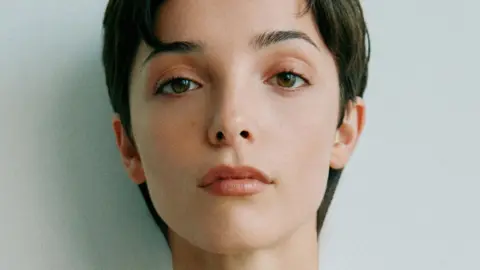 Jessica Reynolds, a woman with short, dark hair, looks to the camera with a neutral expression for a close-up photo. She has brown eyes and is lightly made up. 