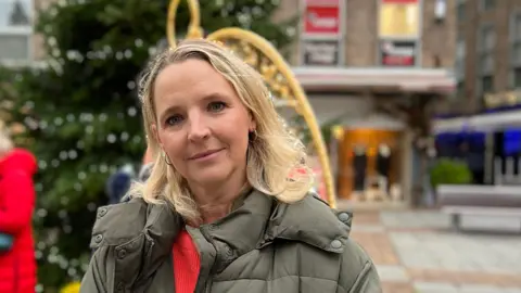 Kate Wright standing in St Helier. She is wearing a green coat and red jumper and looking into the camera.