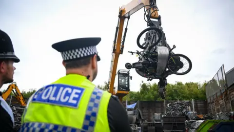 Two police officers watch as a scrap yard telehandler carries an assortment of e-bikes and scooters to a crusher