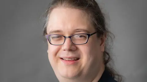 Headshot of Chris Hilliard  - they are wearing a black jacket and black rimmed glasses with long hair in a ponytail