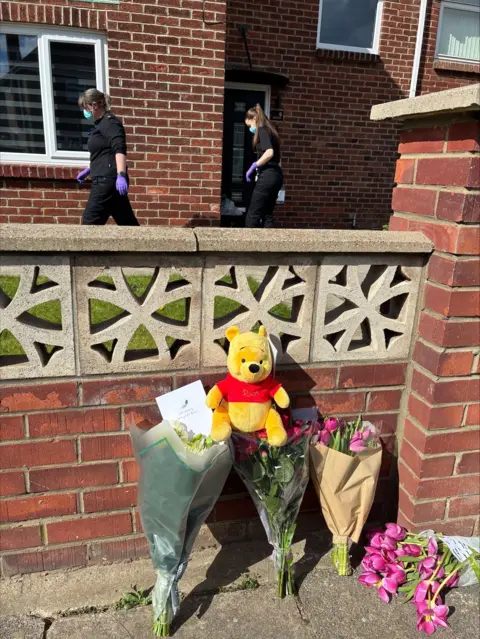 The flowers and Winnie the Pooh toy from the front. Behind the wall, two female officers walk into the house dressed in black. They are also wearing purple gloves and blue face masks.