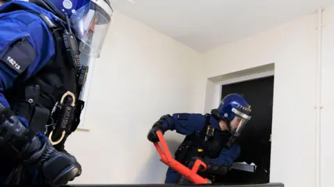 Police handout Police officers wearing riot gear force open a front door with a large metal instrument during a raid.