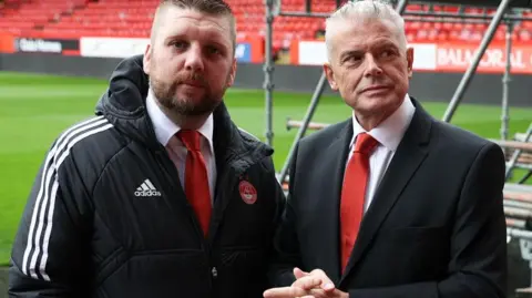 SNS Aberdeen's Alan Burrows and Dave Cormack at Pittodrie. Cormack (left) is wearing a black suit and red tie. Burrows is in a red tie and branded Aberdeen FC jacket. 