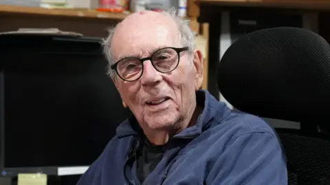 David is smiling in his home office, sitting in an office chair. He has grey hair is wearing a navy pullover and black-rimmed reading glasses.