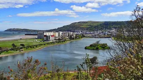 MANX SCenes An arial view of Mooragh Park,  you can see the large lake with a small island in between, there are buildings in the background, there is a hill in the background and you can see a beach on the side.