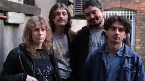 Four members of Brown Horse standing together and looking at the camera outdoors in front of brick and concrete walls.