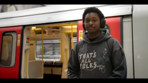 A young black man wearing a green hoodie and headphones looks directly into the camera as he stands on an underground platform. Behind him is a red and white tube with doors open showing folded seats and yellow handrails.