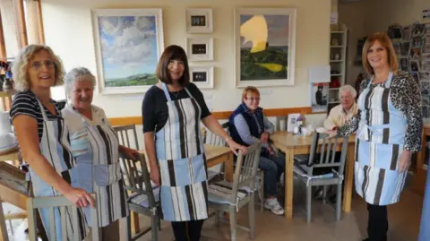 A team of four volunteers in identical stripped blue, black and white aprons, in the cafe, smiling to camera, with two customers at a table in the background.