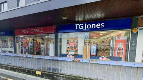 A TG Jones shop with its name in white on blue background with a post office next door. There is a wide pavement in front and blue, rusted railings.