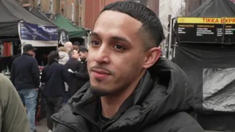 A close-up of a Abdullah being interviewed outdoors at a street food market. He is wearing a black padded jacket and standing in front of food stalls with dark canopies. Other people queue behind him, and a menu board reading “Tikka Express” is visible in the background.