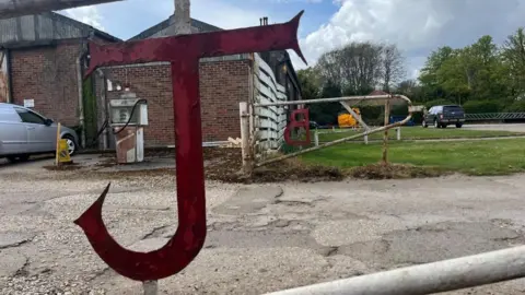A large red J cut out in steel and mounted into a gate. On the opposite side of the driveway, is a symmetrical gate with the letter "B" visible in reverse, denoting Johnson Brothers. There is a white board fence beyond, and the main showroom and workshops, with grass lawns to the right of the image.