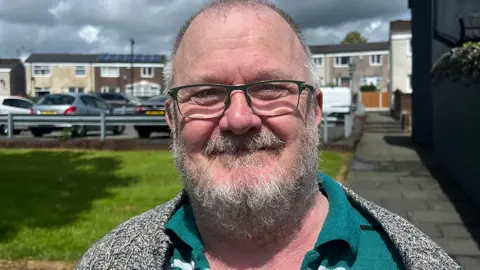 Head and shoulders of David Morley smiling and standing by a grassed area near his home. He has a short grey beard and glasses