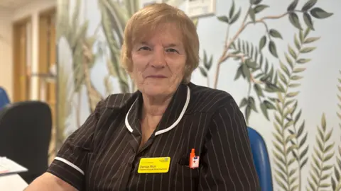 North West Anglia Foundation NHS Trust Denise, a woman, in a brown and white striped nursing uniform with a yellow name badge pinned on it. She is smiling for the camera. She is sitting down on a blue chair, with mostly white-coloured patterned wallpaper behind her.