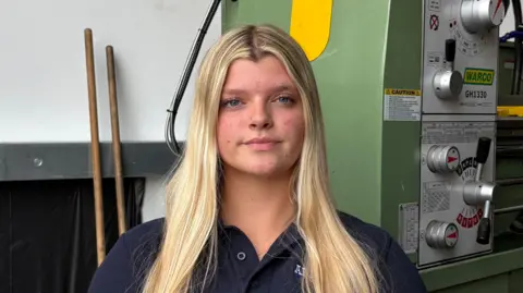 Ben Schofield/BBC Hope Hayden-Ferguson looking straight to camera. She has long blonde hair, which is falling over her shoulders, and blue eyes. She is wearing a dark blue t-shirt and is sitting in front of a large piece of industrial machinery.