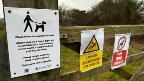 Leanne Brown/BBC Three signs on a wooden gate. A white sign has text that urges dog walkers to follow the Countryside Code. A yellow sign says "Danger - sheer drop off bridge". The third sign is red and says "Keep dogs on leads".