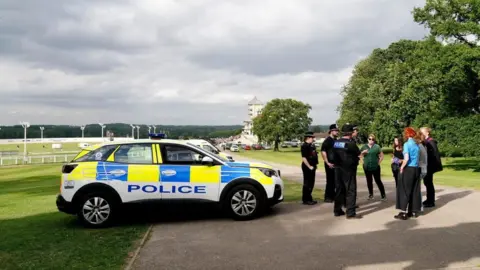 PA Media Police officers stand next to a police car talking to a group of people at a racecourse