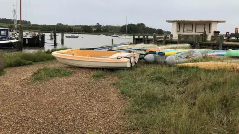 Vikki Irwin/BBC The river Deben from the quayside. It's a slightly grey day and the picture is looking out onto the river. There is a small shingle river bank with boats in the foreground and then a shelter. There are some boats on the river too. 