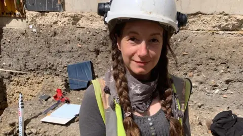 Jess Twyman is standing on another dig on a sunny day. She is wearing a white hard hat and has a green hi-vis jacket and a grey scarf. Her hair is in plaits. The dig is all around her with soil dug out of the site and equipment nearby.