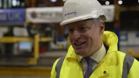 A man wearing a hi-vis jacket and a white hard hat. He is smiling and looking to the side of the camera. He is wearing a white shirt and purple tie under the jacket. He is standing in a boat yard. The background behind him is blurry.