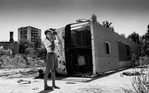 Chris Leslie A child holds a camera next to a destroyed bus