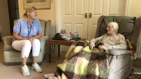 BBC A white woman in a blue top and white trousers sitting in a chair next to an older woman with grey hair and a blanket over her knees