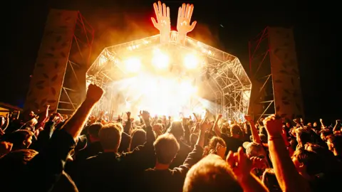 At night a crowd stands in front of the main stage, pumping their arms in the air. The outdoor stage is floodlit so the performers are not really visible, but there is clearly a band on stage. The top of the stage frame has the model of two huge hands pointing upwards.