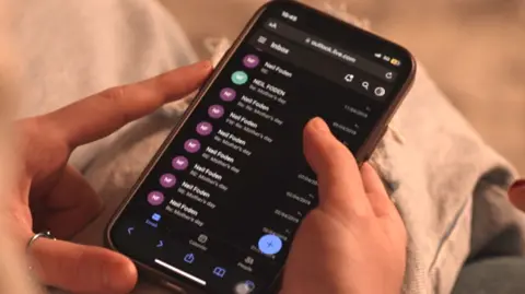 A mobile phone is held in two hands on someone's lap, with a ring on the thumb of the left hand of the person holding it. It shows the Outlook email app open, with a list of messages from Neil Foden in the inbox, most of which are headed 'RE: Mother's Day'.