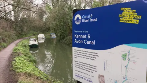 A view of a Canal and River Trust blue and white sigh that says Kennet and Avon Canal. The Canal itself is in the background with several small white boats in the distance.
