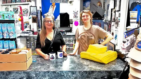 Pallab Ghosh/BBC News Inside a small, crowded NASA gift shop, two people stand behind a grey marble-effect counter. Shelves and walls around them are packed with space souvenirs, mission posters, and astronaut photos. On the left are boxes of mugs; the woman holds two white mugs decorated with NASA-style logos. Next to her, a younger person in a pale T‑shirt leans on the counter near two neat piles of bright yellow folded T‑shirts. To the right, a rack displays beige and orange NASA baseball caps and small astronaut toys, giving the scene a busy, colourful, fan-filled atmosphere.