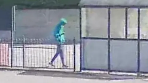 Axel Rudakubana, in a green hoodie and blue face mask, walks on a street near a bus stop