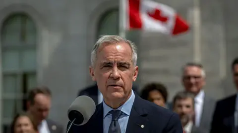 Man with suit and tie on stands in front of microphone while Canadian flag waves in the background