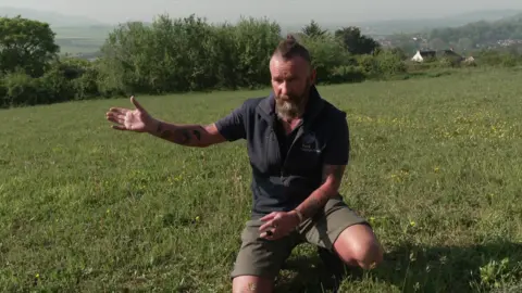 A man in shorts and a gilet points at a field, which he is kneeling in. It is very sunny.