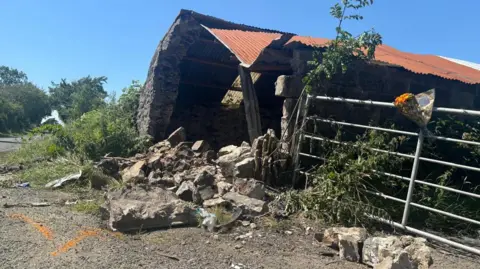There is an old looking shed and wrought iron gate on the right of the picture. There is a lot of rubble at the entrance to the shed, which has a corrugated iron orange roof. There is a road on the left and some flowers have been tied to the gate. There are also some orange arrow markings on the ground in front of the shed.