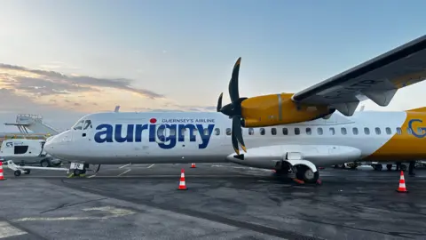 A white and yellow aurigny ATR aircraft on the tarmac in Guernsey. There are cones around the plane.