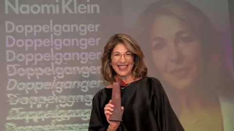 Getty Images Naomi Klein, wearing a black silk top, holding her award for best non-fiction