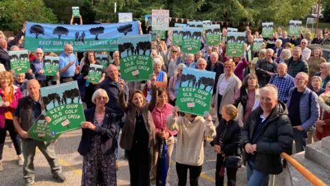 People gathering looking up a camera holding lots of signs saying 'keep our odl golf course green'